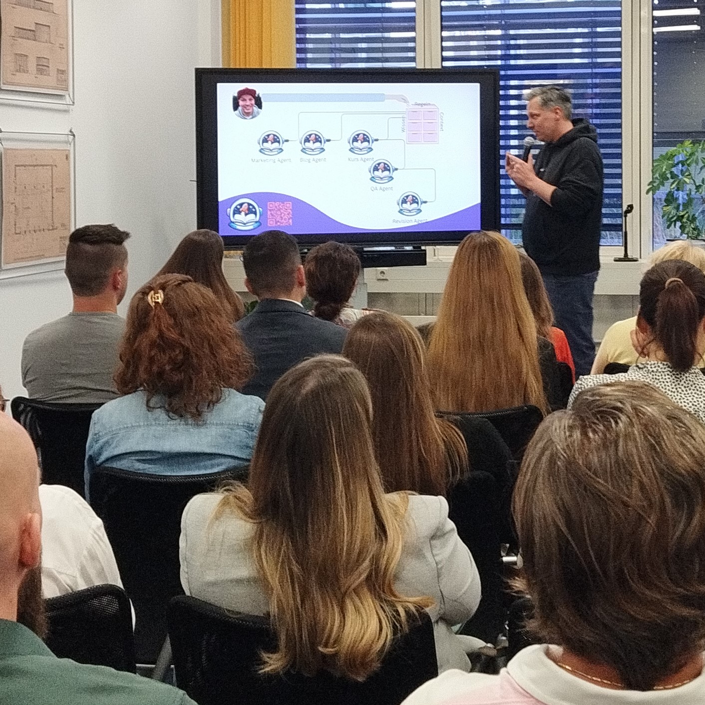 Blick in den Vortragsraum mit Monitor und Referenten, das Publikum ist von hinten zu sehen